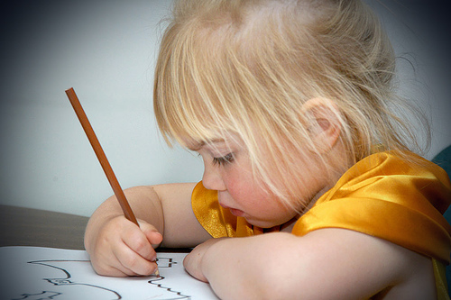 A blonde girl uses a brown colored pencil to color in an image of a bat