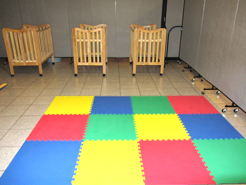 3 cribs lay next to an area of colorful foam square floor tiles arranged for infant play