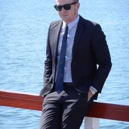 Man in a blue suit and dark sunglasses leans against the rail of a ship.
