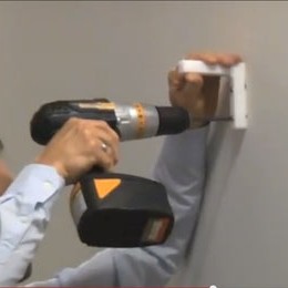 A man uses an electric screwdriver to attach an L bracket to a wall.